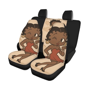 May include: Car seat covers featuring a cartoon character in a red dress, gold earrings, and a white headpiece. The seats have a tan background with heart shapes and black accents.