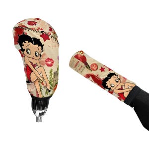 May include: A gear shift knob cover and a handbrake cover featuring a cream-colored background with a repeating print of a cartoon character in a red dress, red lipstick kisses, red stars, and text including the word London in black ink.