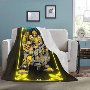 Puede incluir: Una manta de forro polar negra y amarilla con un personaje de robot amarillo en el centro. El robot está de pie con los brazos levantados y tiene un diseño amarillo y negro. El texto "Bumblebee" está en la parte superior de la manta.