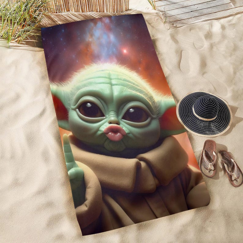 Puede incluir: Una toalla de playa con una ilustraci&oacute;n digital de un personaje verde con aspecto de extraterrestre con ojos grandes y una mueca. La toalla est&aacute; sobre arena, con un sombrero negro y sandalias cerca.