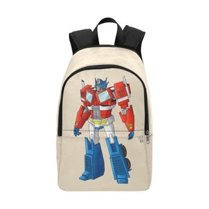 May include: Beige backpack featuring a colorful illustration of Optimus Prime from Transformers. The backpack has black straps and a zipper closure. The Optimus Prime graphic is red, blue, white, and yellow.