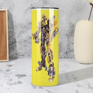 May include: A yellow tumbler featuring a detailed illustration of a robot character in shades of yellow, black, and gray. The tumbler has a silver lid and is designed to hold beverages. The background is a solid yellow color.