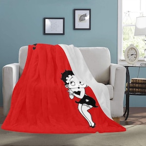 Op de afbeelding: Een rode en witte deken met een cartoon afbeelding van Betty Boop in een zwarte jurk. De deken heeft een zachte, pluche textuur.