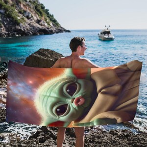 Puede incluir: Una toalla de playa con un primer plano de un personaje alien&iacute;gena verde con ojos grandes y una peque&ntilde;a lengua rosada. El fondo de la toalla muestra una nebulosa con tonos rojos y naranjas. La persona que sostiene la toalla est&aacute; de pie en una playa rocosa.