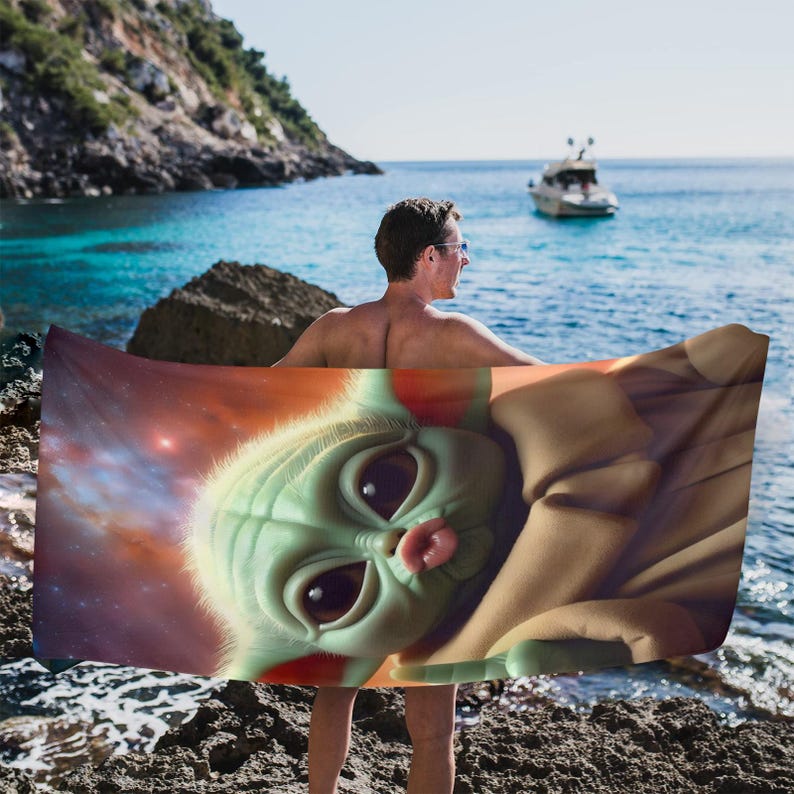 Puede incluir: Una toalla de playa con un primer plano de un personaje alien&iacute;gena verde con ojos grandes y una peque&ntilde;a lengua rosada. El fondo de la toalla muestra una nebulosa con tonos rojos y naranjas. La persona que sostiene la toalla est&aacute; de pie en una playa rocosa.
