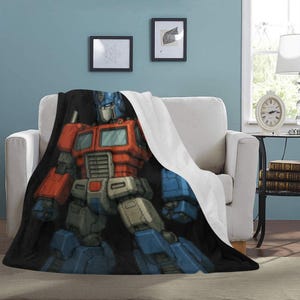 May include: A plush blanket featuring a vibrant illustration of Optimus Prime from Transformers. The blanket showcases the character in his signature red, blue, and silver color scheme. The blanket is draped over a white armchair, with a black background.