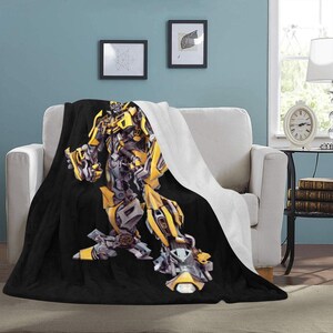 Puede incluir: Una manta de tiro negra y amarilla con una gran imagen de Bumblebee, un robot de la franquicia Transformers. La manta está hecha de tela suave y felpa y es perfecta para agregar un toque de cultura pop a la decoración de tu hogar.