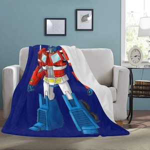 Puede incluir: Una manta de felpa azul y roja con una imagen de dibujos animados de Optimus Prime, un robot de la franquicia Transformers.