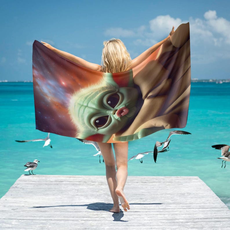 Puede incluir: Una toalla de playa con una imagen grande de un personaje alien&iacute;gena verde con ojos grandes y una boca peque&ntilde;a rosa. La toalla se sostiene contra un fondo de oc&eacute;ano azul con gaviotas volando.