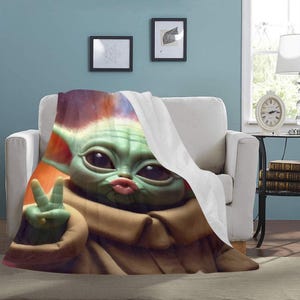 May include: A plush blanket featuring a large graphic of a green alien character with large eyes, puckered lips, and a peace sign. The blanket is draped over a white armchair, with a portion of the blanket folded over to reveal a white underside.
