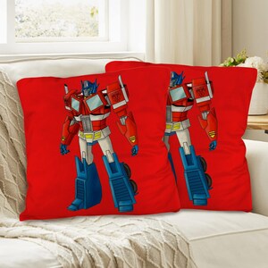 Puede incluir: Dos cojines rojos con una ilustración de dibujos animados de Optimus Prime. El personaje está representado en rojo, azul, blanco y amarillo, sobre un fondo rojo liso. Los cojines están colocados sobre un sofá blanco.