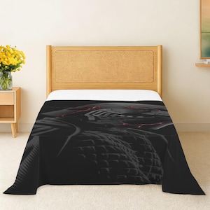 May include: A black and gray blanket with a geometric design and red accents is draped over a bed. The blanket's texture is visible, and the bed has a light wood headboard. The room has a neutral color palette.