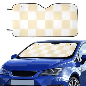 May include: A car sunshade with a checkered pattern in cream and white. The sunshade is rectangular with a black border and eyelets for attachment. The sunshade is shown installed on the windshield of a blue car.
