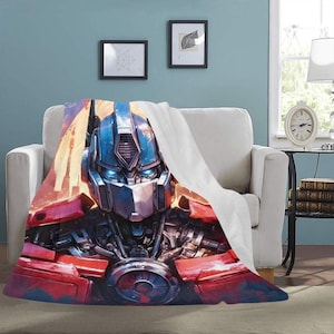 Puede incluir: Una manta de felpa con una ilustración detallada de la cara de Optimus Prime. La manta muestra una vibrante paleta de colores rojo, azul y plateado, con un telón de fondo de paisaje urbano. La manta está sobre un sillón blanco.