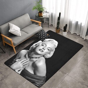 Puede incluir: Alfombra rectangular negra con una imagen en blanco y negro de Marilyn Monroe. La alfombra muestra un primer plano de la actriz con los ojos cerrados, luciendo un collar y un vestido oscuro. La alfombra está colocada sobre un suelo embaldosado.