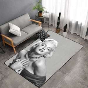 Puede incluir: Una alfombra rectangular con una imagen en blanco y negro de Marilyn Monroe. La alfombra tiene un fondo gris y un borde negro. La imagen muestra a Monroe con los ojos cerrados, usando un collar y un vestido oscuro.