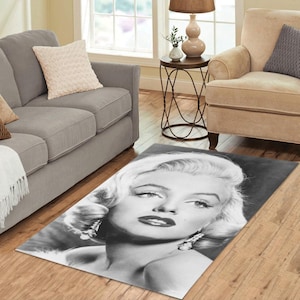 Puede incluir: Alfombra rectangular con un retrato en blanco y negro de Marilyn Monroe. La imagen muestra su rostro con su cabello rubio, labios oscuros y pendientes colgantes. La alfombra está colocada sobre un suelo de madera.