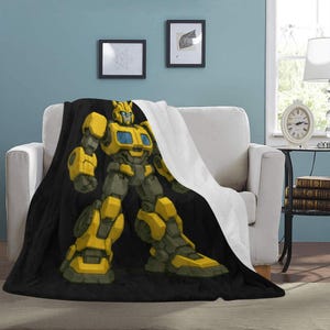 Puede incluir: Una manta en blanco y negro sobre un sofá blanco, con una gran ilustración de un personaje de robot amarillo y gris. El robot está en una pose dinámica, con un diseño detallado. La manta es de material suave.