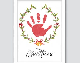 Puede incluir: Impresión enmarcada con una huella de mano roja dentro de una corona verde y roja, adornada con lazos rojos y campanas doradas. Las palabras "Merry Christmas" están escritas en elegante caligrafía debajo.