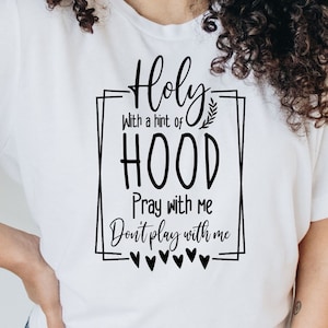 May include: White t-shirt with a black graphic design that says "Holy with a hint of HOOD Pray with me Don't play with me".