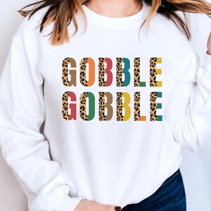 Op de afbeelding: Witte trui met de tekst "GOBBLE GOBBLE" bedrukt in een kleurrijk luipaardmotief.