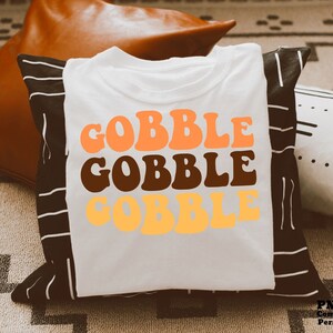 Puede incluir: Camiseta blanca con el texto "GOBBLE GOBBLE GOBBLE" en letras naranja, marrón y amarillo. Las letras están en un estilo de fuente retro.