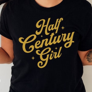 May include: Black t-shirt with gold lettering that reads "Half Century Girl" with small stars around the text.