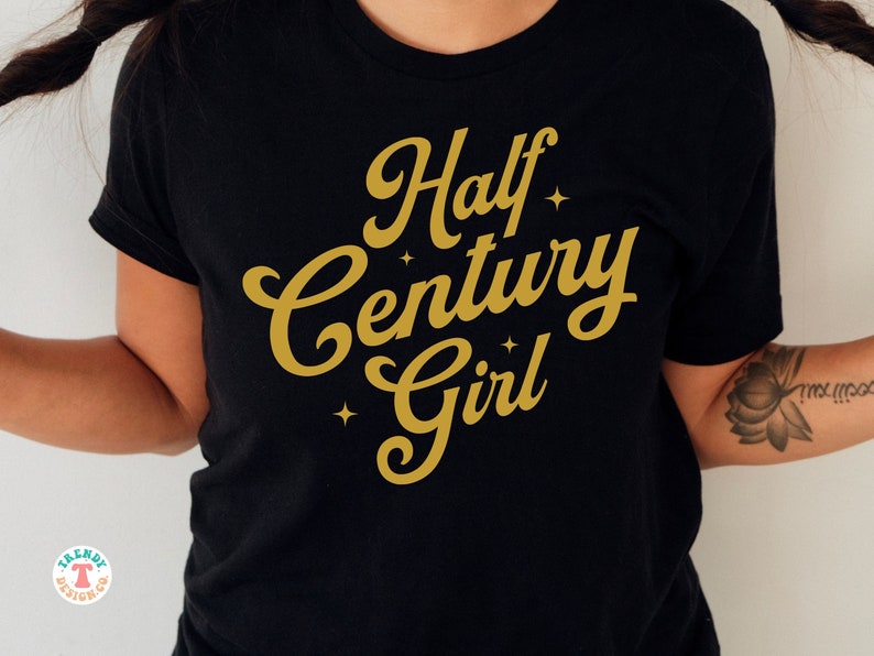 May include: Black t-shirt with gold lettering that reads "Half Century Girl" with small stars around the text.