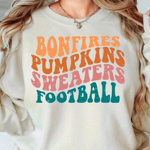 Puede incluir: Una sudadera beige con un diseño gráfico retro que dice "Bonfires, Pumpkins, Sweaters, Football" en letras coloridas y desgastadas.