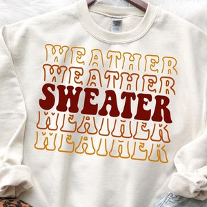 Puede incluir: Una sudadera de color crema con el texto "WEATHER SWEATER" en letras rojas y naranjas.