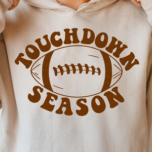 May include: A brown hoodie with the text "Touchdown Season" in a retro font. A brown football graphic is centered in the text.