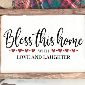 Op de afbeelding: Een wit houten bord met de tekst "Bless this home with love and laughter" in zwarte cursieve letters. Er zijn kleine rode hartjes boven en onder de tekst.