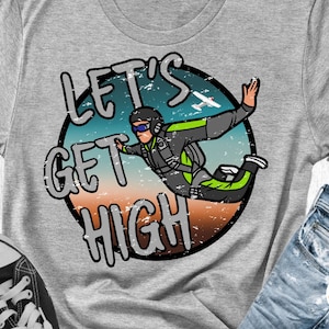 Puede incluir: Una camiseta gris con un gráfico circular desgastado que presenta un paracaidista en el aire con un pequeño avión en el fondo. El texto "Let's Get High" está escrito en una fuente desgastada alrededor del gráfico.