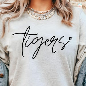 Puede incluir: Una camiseta blanca con la palabra "tigers" escrita en cursiva negra con un pequeño corazón al lado.
