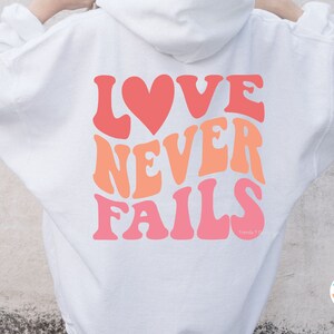 Op de afbeelding: Witte hoodie met een roze, oranje en rode afbeelding met de tekst "LOVE NEVER FAILS" in een retro-lettertype. De afbeelding bevat een hart.