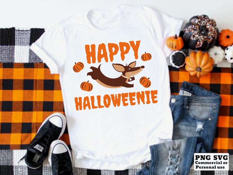 May include: White t-shirt with the text "Happy Halloweenie" in black and orange. A cartoon dachshund wearing a red collar is jumping over a pile of orange pumpkins.