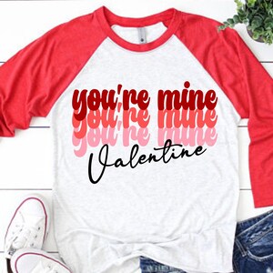 May include: A white and red baseball tee shirt with the text "you're mine" repeated in a layered, retro style with the word "Valentine" underneath.