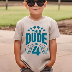 May include: A light gray t-shirt with a blue graphic that says "This Dude is 4" with thumbs up on either side of the number 4. There are five blue stars above the text.