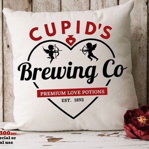 以下が含まれることがあります： ハート型のデザインの中に、黒い筆記体で「CUPID'S Brewing Co」と書かれた白い正方形のクッション。デザインには、弓と矢を持った2人のケルビムが含まれています。「PREMIUM LOVE POTIONS」と「EST. 1892」の文字も赤色で表示されています。
