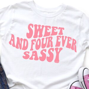 May include: White t-shirt with pink text that reads "Sweet and Four Ever Sassy" in a retro font.