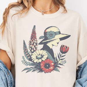 May include: Ivory-colored t-shirt featuring a floral design with a woman in a hat. The graphic includes red, white, and blue flowers, and the text "mind & harmony". The shirt has short sleeves and a relaxed fit.