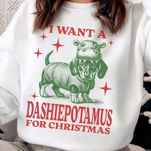 May include: White sweatshirt featuring a red, green and black illustration of a dachshund wearing a hippopotamus costume. The text "I want a dashiepotamus for Christmas" is printed on the sweatshirt.