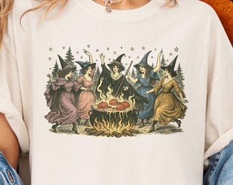 Peut inclure: T-shirt ivoire Comfort Colors avec une illustration vintage de sorcières autour d'un chaudron. Le t-shirt a des manches courtes et un col rond. Le texte "MIND & HARMONY GIFTS" est sur le côté droit de l'image.