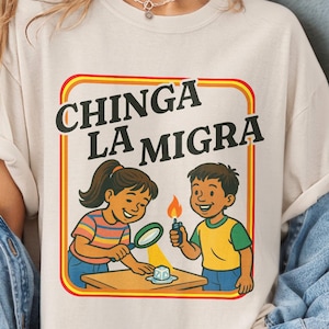 May include: Ivory Comfort Colors t-shirt with a graphic featuring two cartoon children and the text "CHINGA LA MIGRA" in a yellow and orange frame. The shirt also has text that says "MORE COLORS & PRODUCTS AVAILABLE".