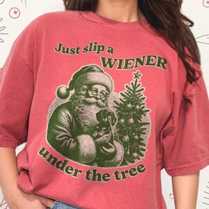 May include: Red t-shirt featuring a vintage illustration of Santa Claus holding a dachshund and a Christmas tree. The text on the shirt reads "Just slip a WIENER under the tree".