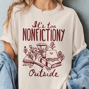 May include: Ivory t-shirt featuring the burgundy text "It's too NONFICTIONY Outside" with book and floral illustrations. Part of the Comfort Colors 1717 range. The shirt is designed to be worn oversized, suggesting sizing up.