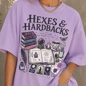 May include: A light purple t-shirt featuring the text "HEXES & HARDBACKS" and "reading spells one chapter at a time" above a graphic illustration of stacked books, a crystal ball, a candle, a teacup, an open spellbook, tarot cards, and ink.