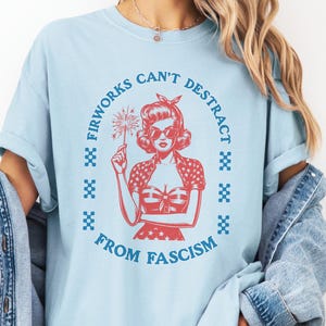 May include: Pale blue t-shirt featuring a red graphic of a woman with a firework, encircled by the words "Fireworks Can't Distract From Fascism." The word "CHAMBRAY" is at the top left.