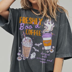 May include: Dark grey t-shirt with a Halloween graphic. The design includes the text "FRESHLY BOO'd COFFEE" in orange and purple, alongside illustrations of coffee cups, ghosts, bats, and eyeballs. The phrase "Served with screams and sugar" is also present.