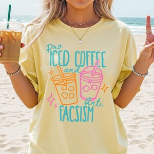 May include: A pale yellow t-shirt with the slogan "Pro Iced Coffee and Anti Fascism" in turquoise and orange. The design includes illustrations of iced coffee cups. The person is holding an iced coffee drink.